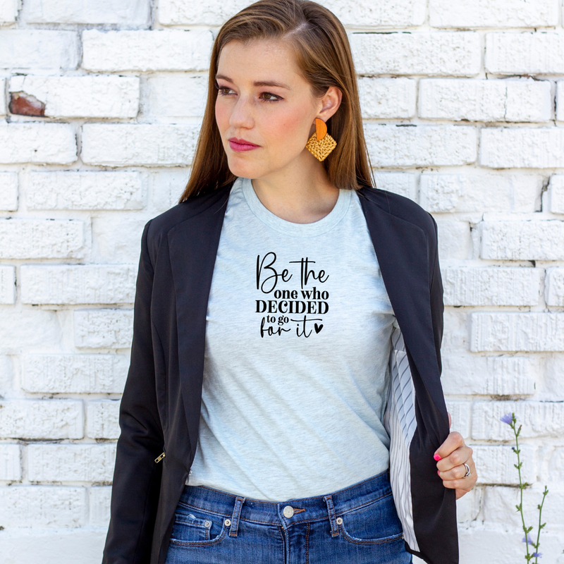 A woman wearing a Bella Canvas heather ice blue t-shirt with a motivational quote, "Be the one who decided to go for it", standing against a white brick wall