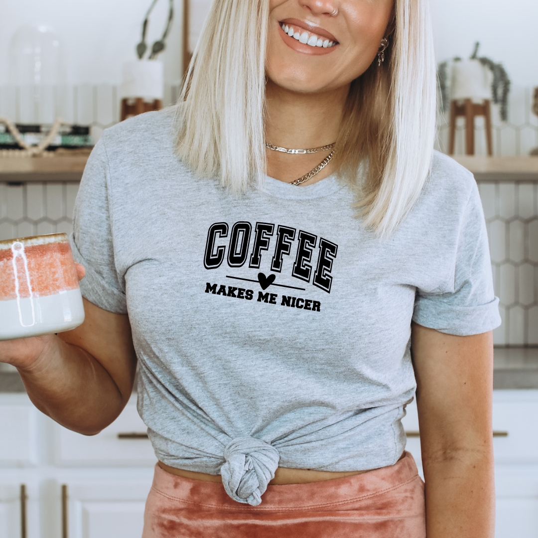 Woman wearing a Bella Canvas heather gray graphic t-shirt with the phrase "Coffee Makes Me Nicer" while holding a coffee mug in a bright kitchen.