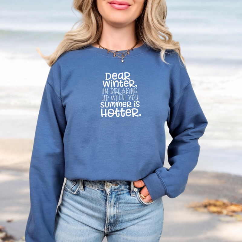Woman standing near the beach wearing a Gildan Indigo Blue sweatshirt with white lettering that reads “Dear Winter, I’m Breaking Up With You Summer Is Hotter.” with her hand in her jeans pocket.