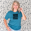 Woman wearing a deep heather teal Bella Canvas t-shirt with black retro lettering that reads “Grow Positive Thoughts” surrounded by flower illustrations, standing in front of a spotted background.