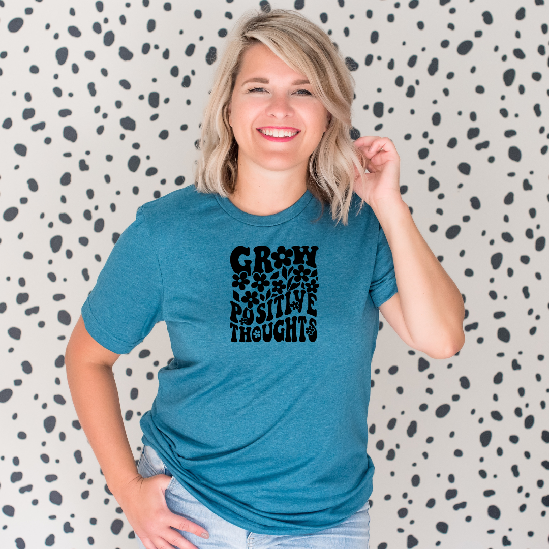 Woman wearing a deep heather teal Bella Canvas t-shirt with black retro lettering that reads “Grow Positive Thoughts” surrounded by flower illustrations, standing in front of a spotted background.
