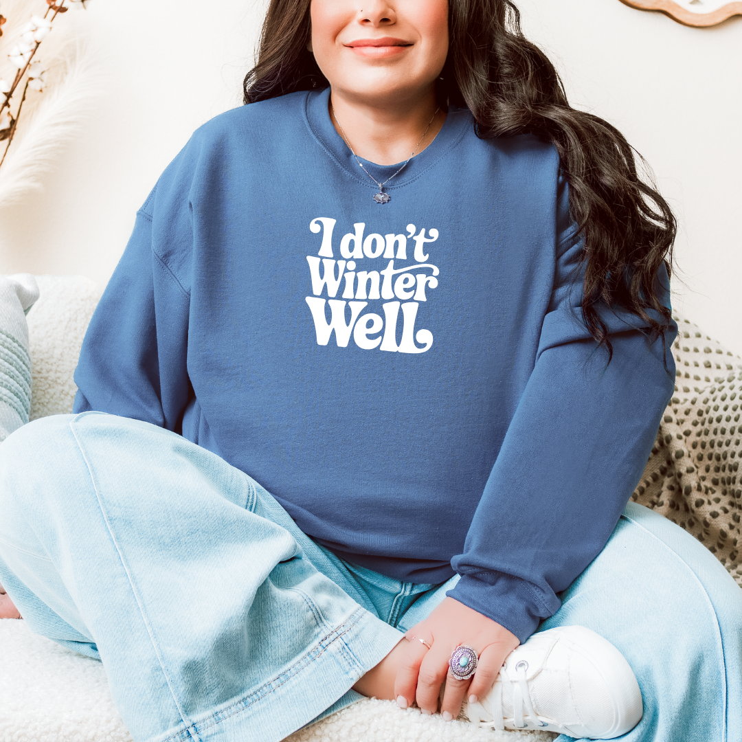 Woman sitting cross-legged on a couch wearing a Gildan Indigo Blue sweatshirt with white retro lettering that reads “I Don’t Winter Well,” smiling softly.