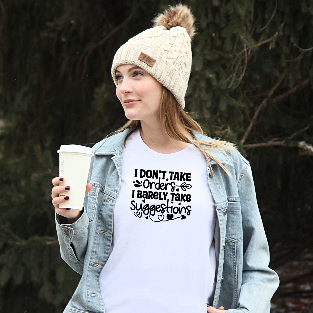 Woman wearing a Bella Canvas white graphic t-shirt with the phrase "I Don't Take Orders, I Barely Take Suggestions" under a denim jacket while holding a to-go coffee outdoors.