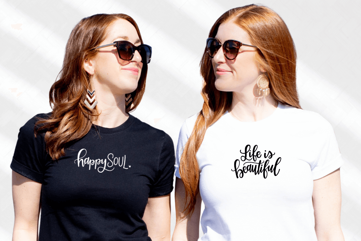 Two women are looking at one another. The woman on the left is wearing a Bella Canvas heather black tee with the white script "happy soul." The woman on the right is in a white Bella Canvas tee with black script "Life is Beautiful."