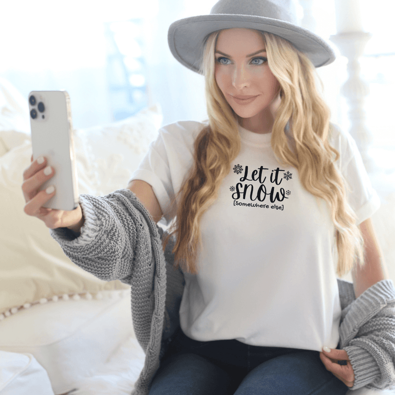 Blonde woman taking a selfie wearing a white bella canvas 3001 tee with snowflakes and let it snow somewhere else in black lettering - a moxie momma original design
