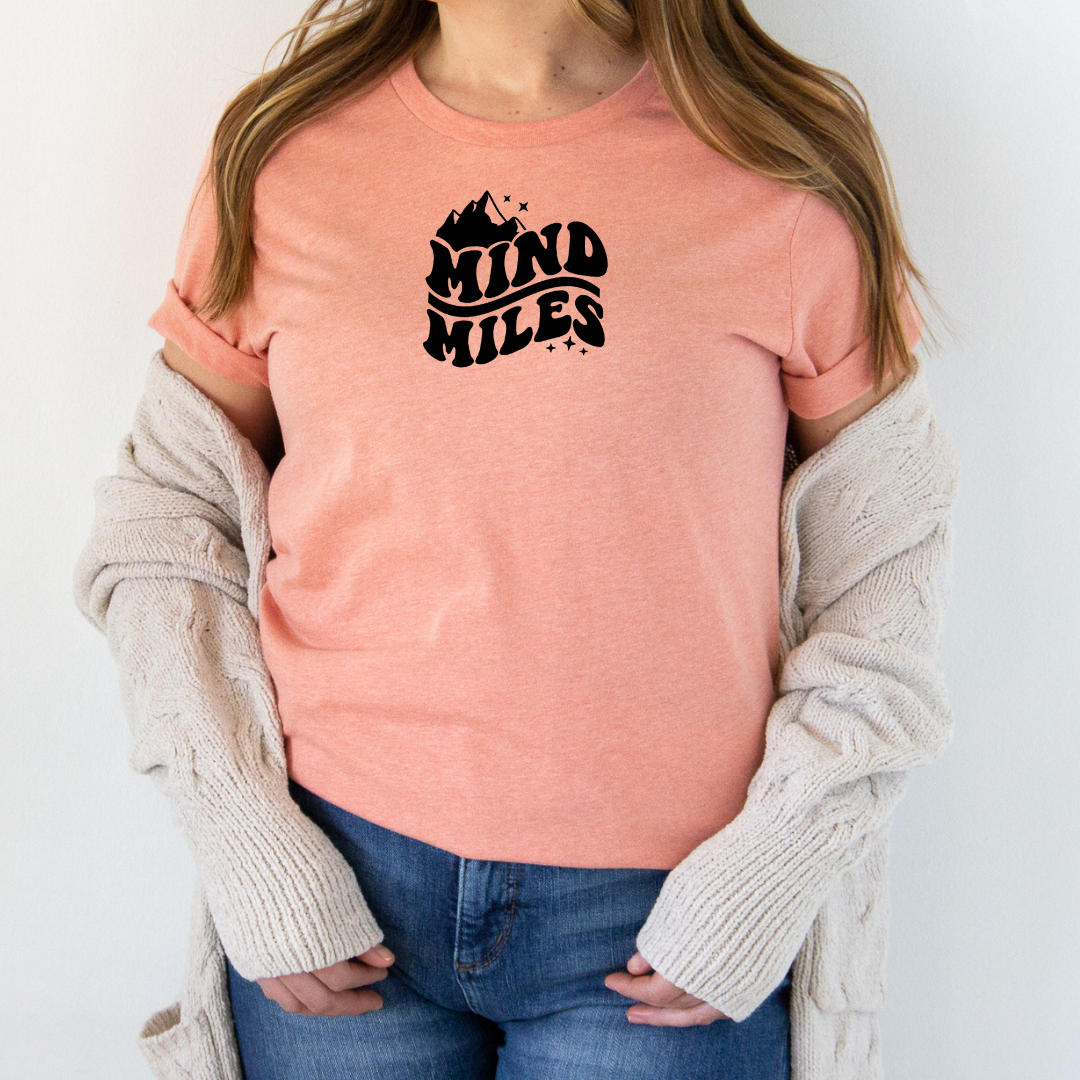 Person wearing a Bella Canvas heather sunset t-shirt with 'Mind Miles' text, with a mountain graphic, standing against a plain background