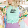 Woman wearing a heather mint green Bella Canvas t-shirt with black retro lettering that reads “Sunshine Mixed with a Little Hurricane,” styled with a colorful floral kimono and purple leggings.