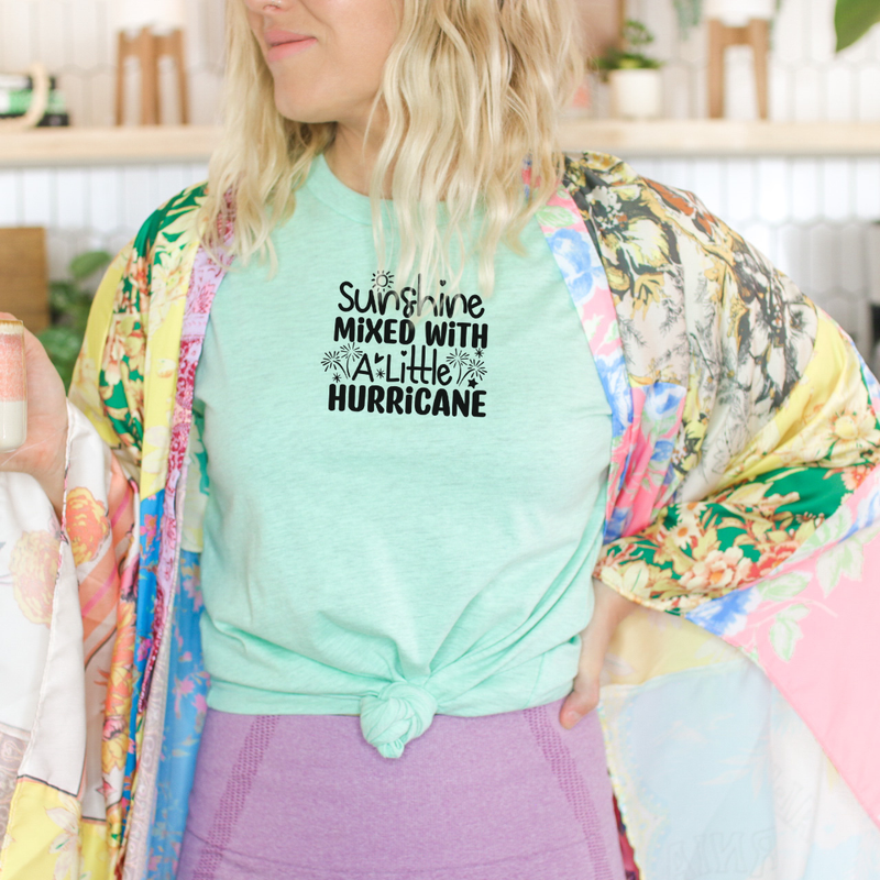 Woman wearing a heather mint green Bella Canvas t-shirt with black retro lettering that reads “Sunshine Mixed with a Little Hurricane,” styled with a colorful floral kimono and purple leggings.