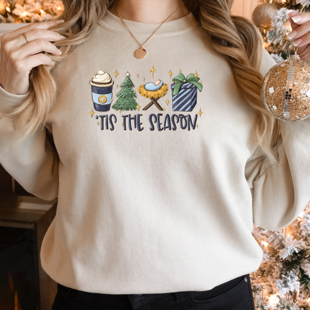 Woman wearing a Sand Gildan 81000 Crewneck Sweatshirt with a DTF image in deep blues, green, and gold with text tis the season. Image includes hot beverage, Christmas Tree, gift with bow, baby Jesus in a manger. 