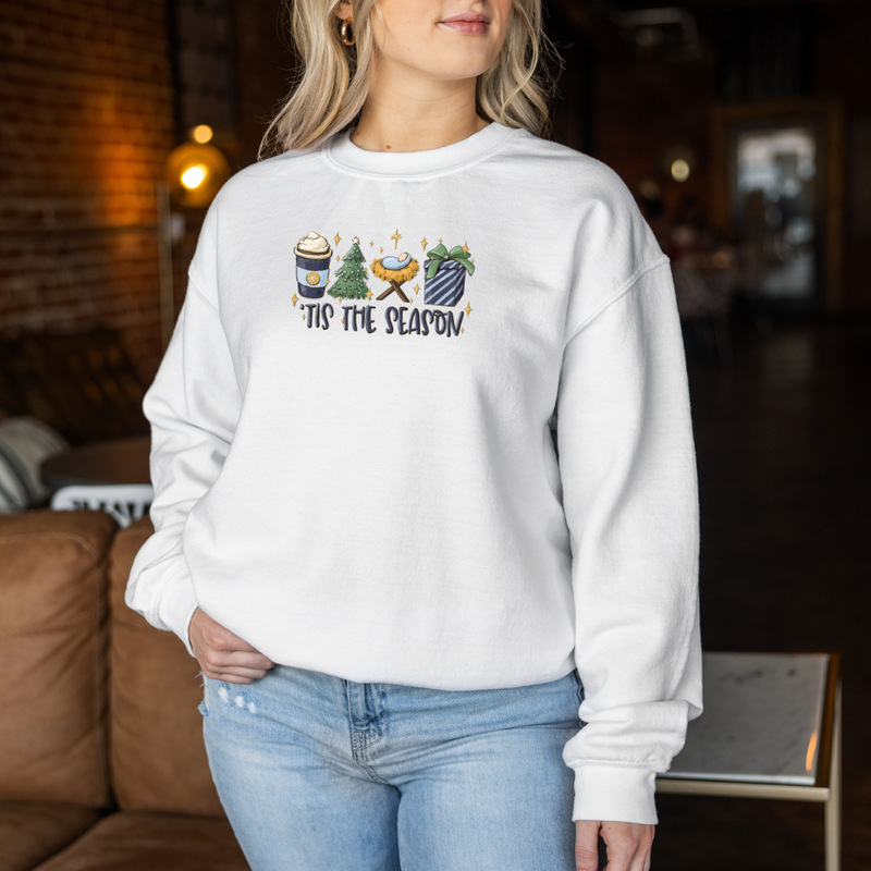 Woman wearing a White Gildan 81000 Crewneck Sweatshirt with a DTF image in deep blues, green, and gold with text tis the season. Image includes hot beverage, Christmas Tree, gift with bow, baby Jesus in a manger. 