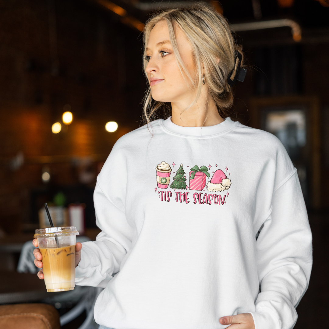 Woman wearing White Gildan 81000 Crewneck Sweatshirt with a DTF image in pink with text tis the season. Image includes hot beverage, Christmas Tree, gift with bow, santa hat.