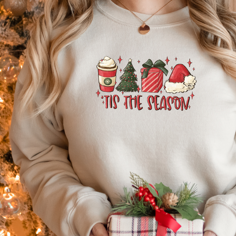 Woman wearing a Sand Gildan 81000 Crewneck Sweatshirt with a DTF image in red with text tis the season. Image includes hot beverage, Christmas Tree, gift with bow, santa hat. 