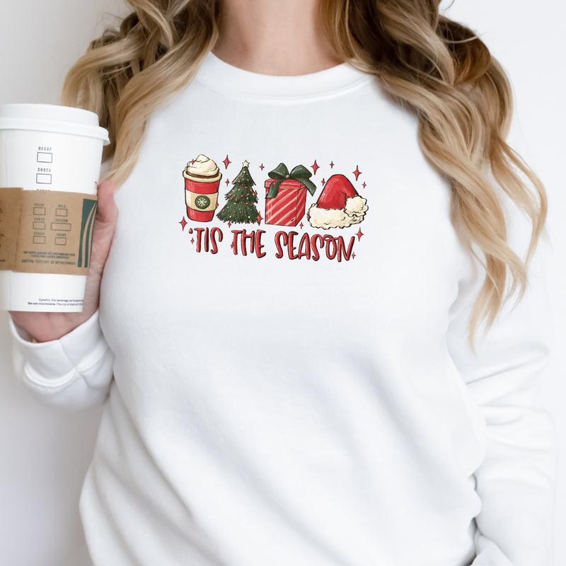 Woman wearing a White Gildan 81000 Crewneck Sweatshirt with a DTF image in red with text tis the season. Image includes hot beverage, Christmas Tree, gift with bow, santa hat. 