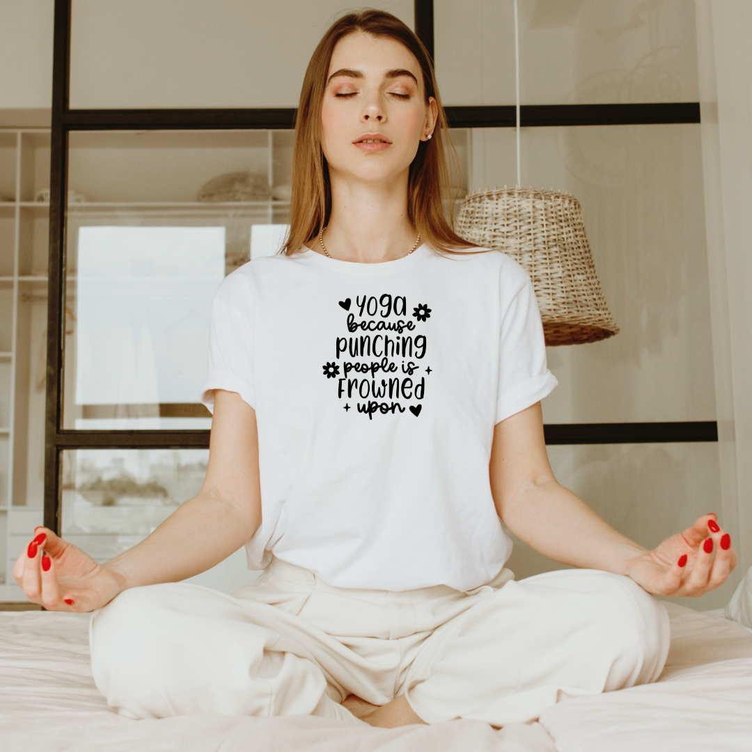 Woman sitting cross-legged in a meditation pose wearing a Bella Canvas white t-shirt with black lettering that reads "Yoga Because Punching People Is Frowned Upon."
