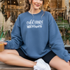 Woman sitting outside a shop wearing a Gildan Indigo Blue sweatshirt with white “Cold Hands Warm Heart” lettering, holding an iced coffee and smiling.