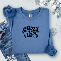 Folded Gildan Indigo Blue crewneck sweatshirt with black “Cozy Vibes” lettering, styled with jeans, a scrunchie, and snowflake decorations for a winter aesthetic.
