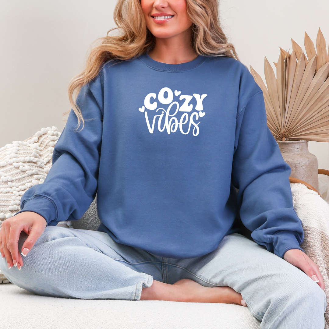 Woman sitting cross-legged wearing a Gildan Indigo Blue crewneck sweatshirt with white lettering that reads “Cozy Vibes,” smiling and relaxed in a soft, neutral room.
