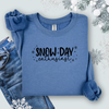 Folded Gildan Indigo  Blue sweatshirt with black lettering that reads “Snow Day Enthusiast,” surrounded by jeans winter-themed decor.