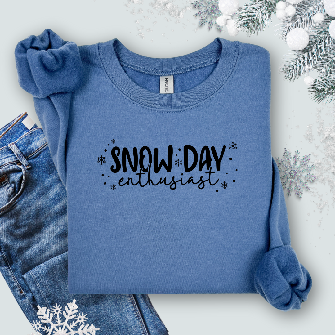 Folded Gildan Indigo  Blue sweatshirt with black lettering that reads “Snow Day Enthusiast,” surrounded by jeans winter-themed decor.