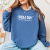 Woman wearing a Gildan Indigo Blue sweatshirt with white “Snow Day Enthusiast” lettering, gently lifting the collar while sitting comfortably in a cozy indoor setting.
