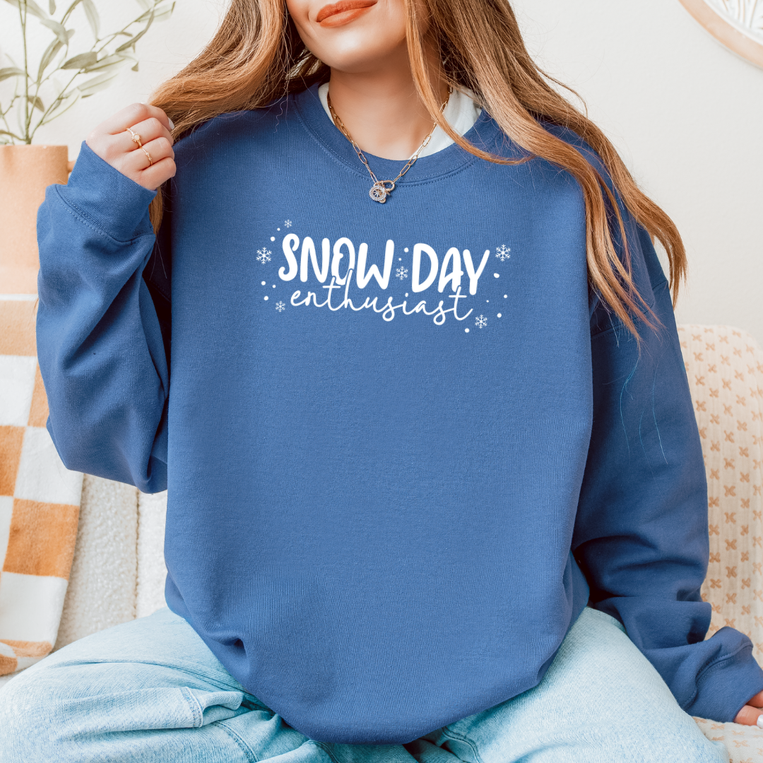 Woman wearing a Gildan Indigo Blue sweatshirt with white “Snow Day Enthusiast” lettering, gently lifting the collar while sitting comfortably in a cozy indoor setting.