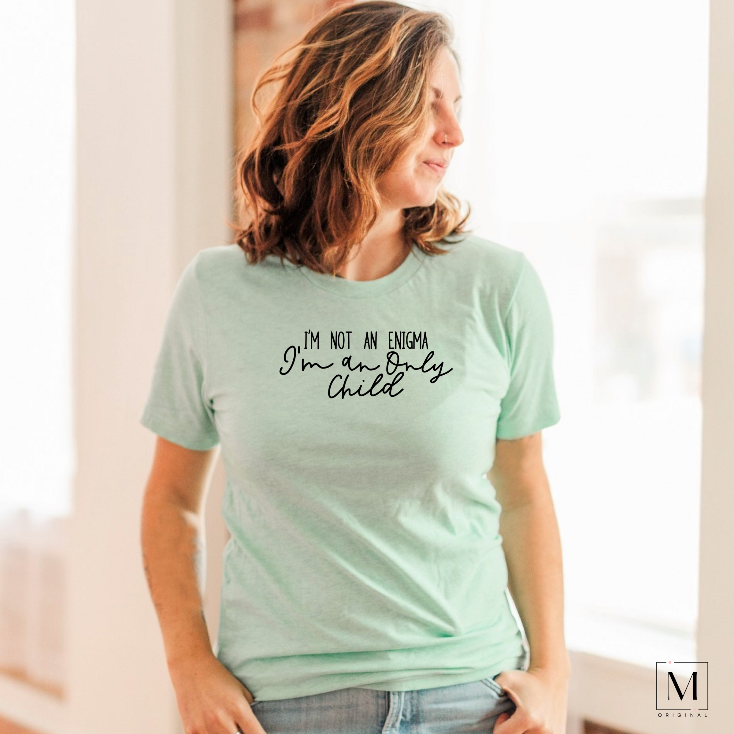 A woman wearing a Bella Canvas 3001cvc heather prism mint t-shirt with black lettering reading I'm not and enigma I'm an only child. Part of Moxie Momma's exclusive only child line.