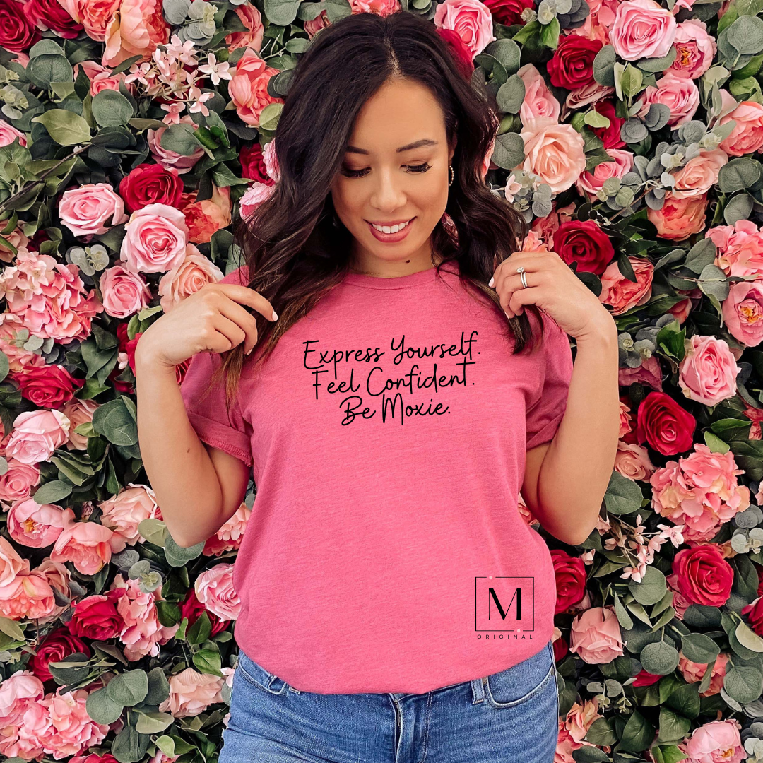 A woman wearing a Bella Canvas Heather Raspberry shirt with black script reading Express Yourself. Feel Confident. Be Moxie. Moxie Momma's tagline and original design.