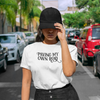 Woman standing in a street between cars wearing a white Bella Canvas 3001 tshirt with black text reading Paving My Own Road. A Moxie Momma Exclusive design.